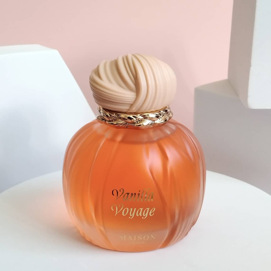 Round perfume bottle with Vanilla Voyage label, a pink cap and gold trim, on a white base